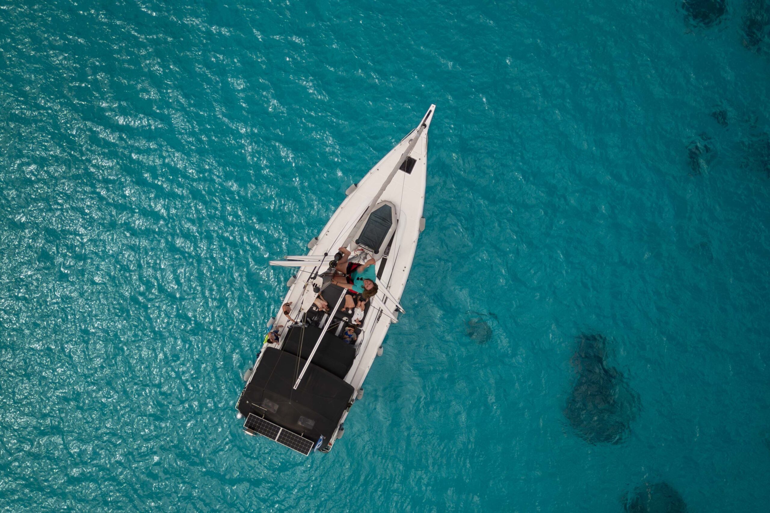 Luftaufnahme eines Segelboots mit einigen Personen an Deck, die während ihrer Segelreise Griechenland in klarem türkisfarbenem Wasser treiben. Der weiße Rumpf des Bootes kontrastiert mit dem leuchtend blauen Meer und schafft eine ruhige und malerische Szene.