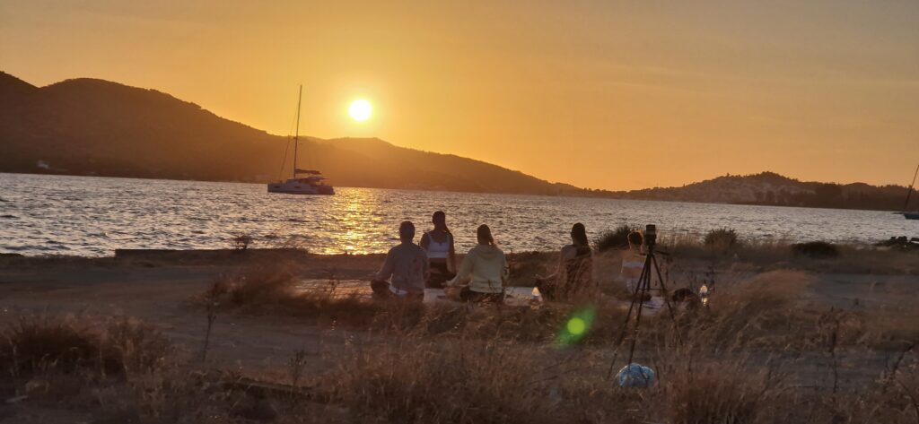 Eine Gruppe von Menschen sitzt auf dem Gras neben einem ruhigen Gewässer und beobachtet den Sonnenuntergang. Ein Boot mit Segel liegt auf dem Wasser, was an Szenen aus dem Skippertraining im Mittelmeer erinnert, und im Hintergrund sind Hügel zu sehen. Der Himmel leuchtet orange und reflektiert den Sonnenuntergang.