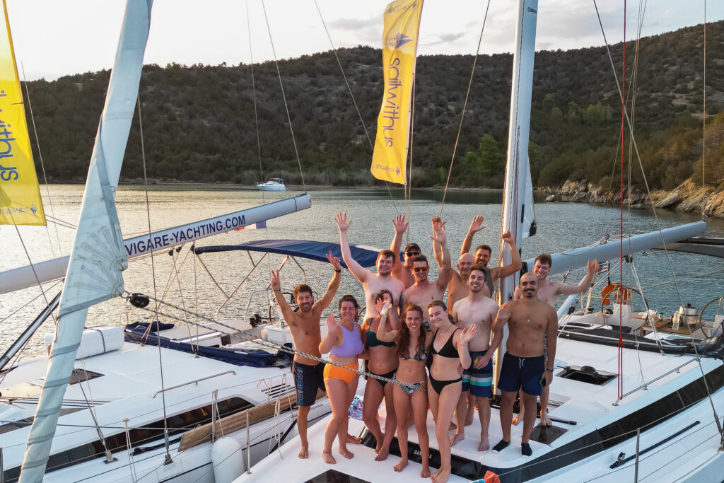 Eine Gruppe von Menschen steht lächelnd und winkend auf dem Deck einer weißen Yacht. Sie tragen Badebekleidung und scheinen einen sonnigen Tag während ihres Segelurlaubs auf Sardinien zu genießen. Neben ihnen ist eine weitere Yacht zu sehen, im Hintergrund sind malerische Hügel und Bäume zu sehen. Tipps für Reiseleiter: So gestalten Sie unvergessliche Gruppenreisen