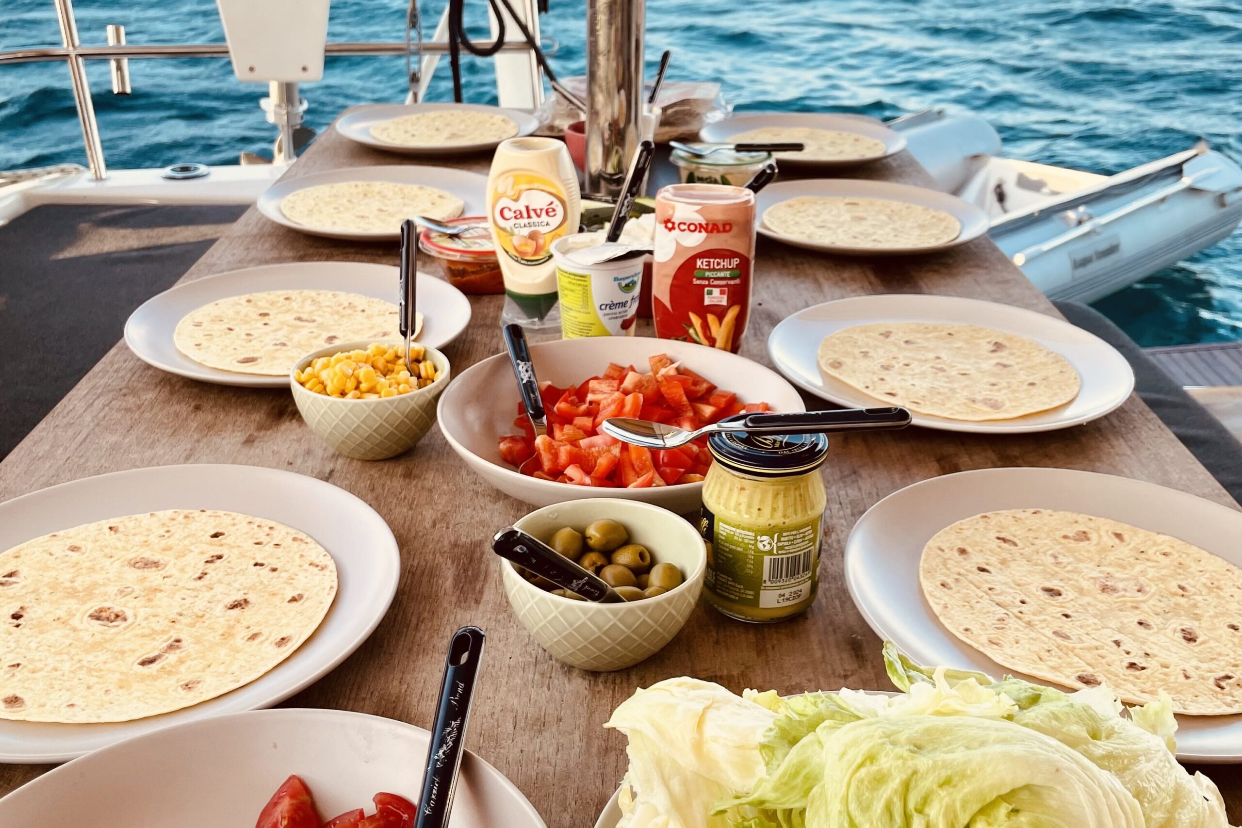 Essen auf einem Segelboot um als Tipp für budgetfreundliches Reisen möglich zu machen auf einem Segeltörn für junge Leute. Man sieht Wraps und Zutaten wir Oliven, Mais, Tomaten, Salat, verschiedne Soßen und das wunderschöne blaue Meer im Hintergrund.