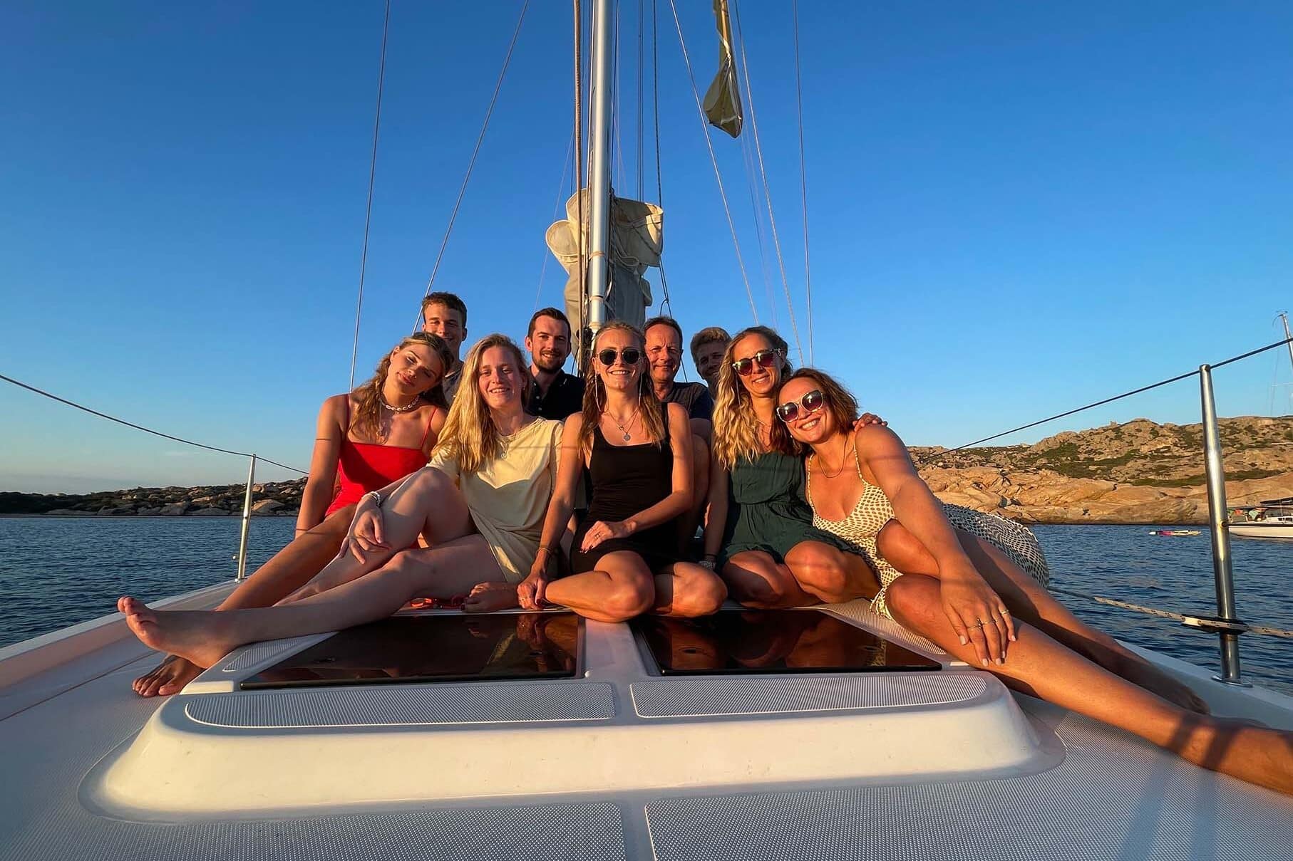 Eine Gruppe von acht Freunden posiert während ihrer Segelreise bei Sonnenuntergang auf dem Vorderdeck eines Segelboots und lächelt freudig vor der malerischen Kulisse des ruhigen Meeres und der felsigen Küste. Kultur