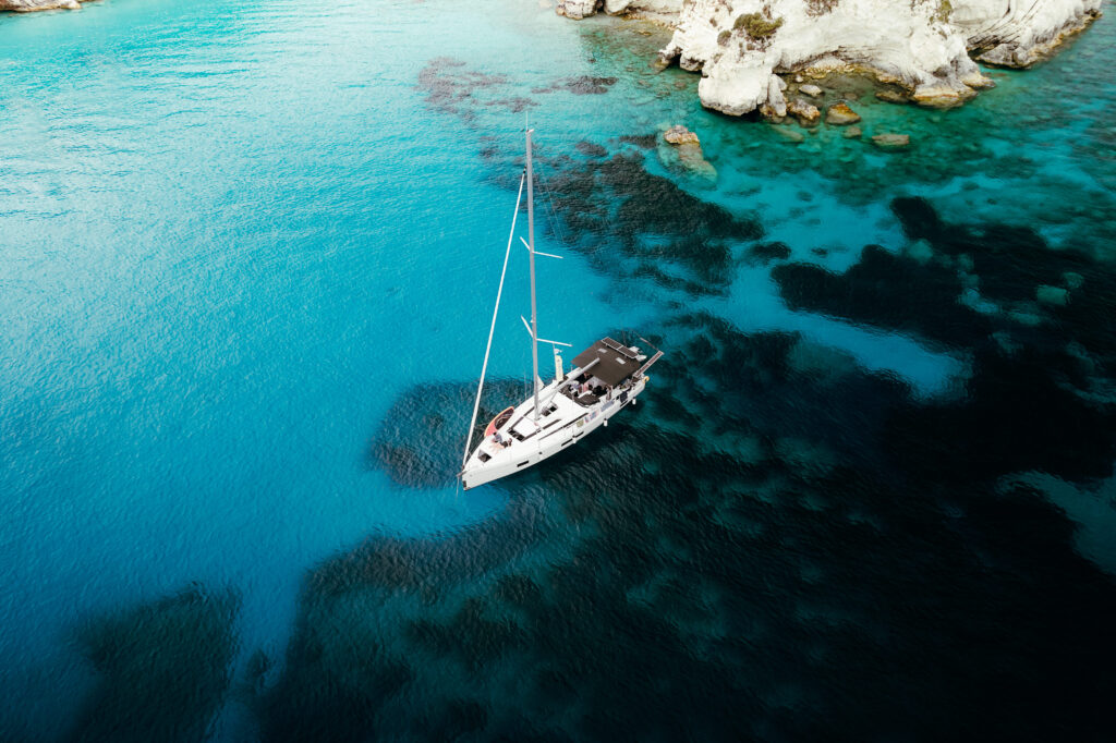 Ein Segelboot ankert in einer ruhigen, türkisfarbenen Bucht, umgeben von teilweise unter Wasser liegenden Felsen und weißen Klippen – perfekt für eine ruhige Pause während einer Atlantiküberquerung. Das Wasser variiert in Blautönen, mit Flecken von dunklerem Blau und Grün um das Boot herum. Kurzurlaub unter Segeln: Die perfekte Auszeit auf dem Wasser
