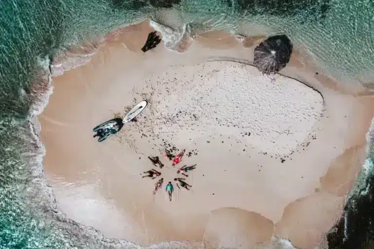 Luftaufnahme eines kleinen, abgeschiedenen Strandes, umgeben von Klippen, mit ein paar Segelbooten und Menschen, die über den Sand verstreut sind, gesäumt von klarem, türkisfarbenem Wasser. Mitsegeln in der Karibik