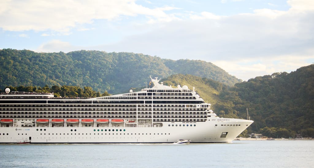 A majestic cruise ship sails through tranquil waters with lush hills in the background, perfect for travel enthusiasts. Mitsegeln vs. Kreuzfahrt: Welche Reiseart passt zu Dir?