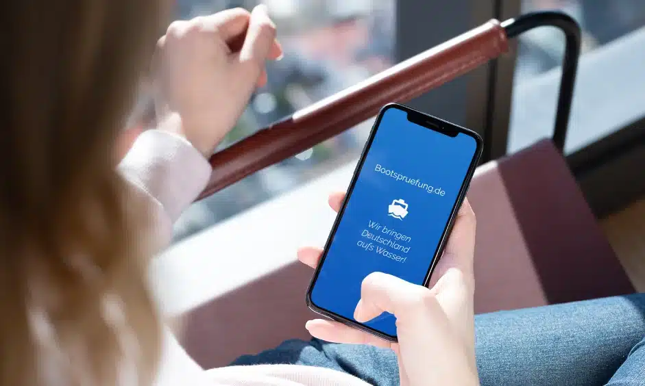 Frau in einem Bus hält ein Smartphone in der Hand, auf dessen Bildschirm eine Buchungsseite für einen Segelurlaub in deutscher Sprache angezeigt wird und die Verfügbarkeit ab heute angibt.