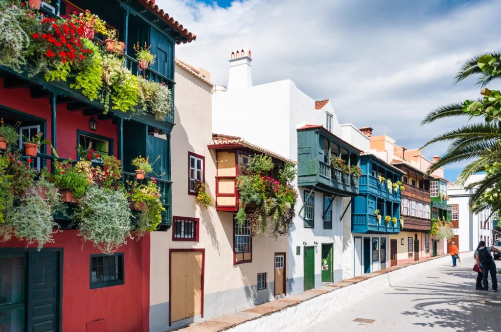 Farbenfrohe, traditionelle spanische Häuser mit Holzbalkonen, die mit leuchtenden Blumen geschmückt sind, säumen unter einem blauen Himmel eine gepflasterte Straße, durch die Menschen spazieren.