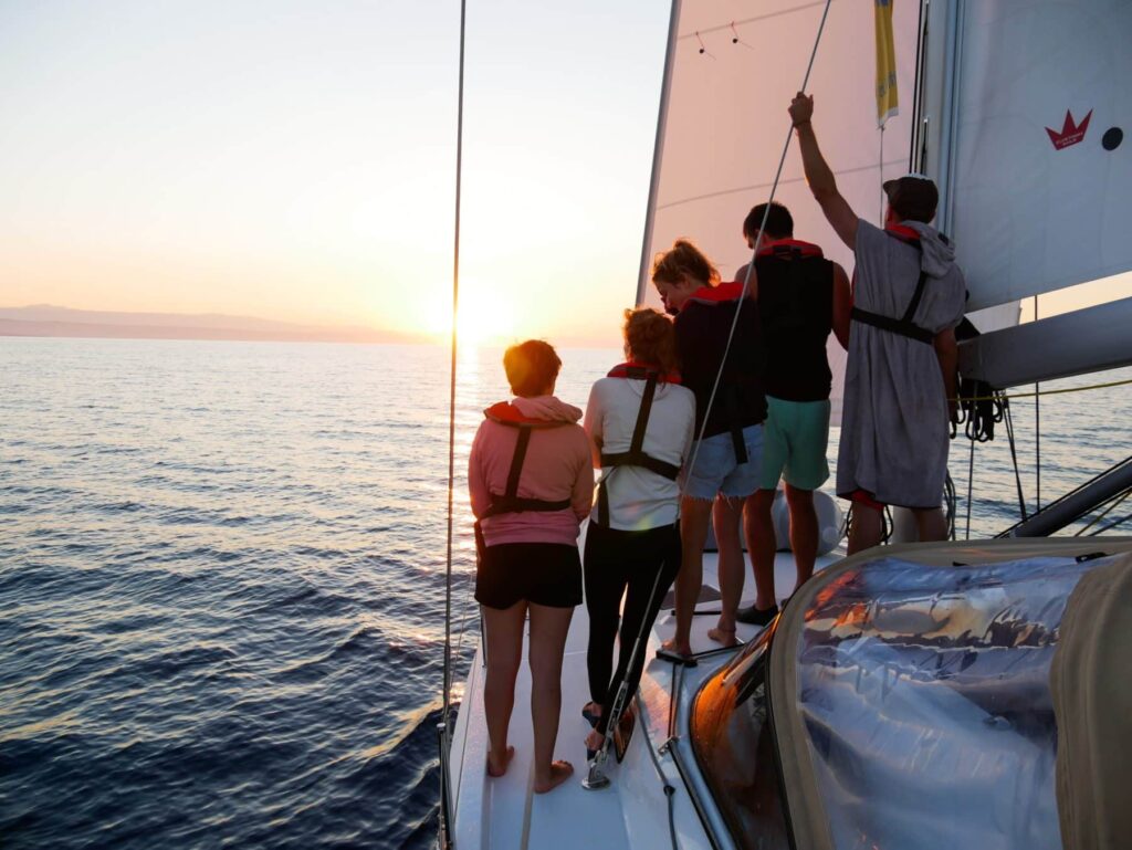 Eine Gruppe von Menschen steht auf einem Segelboot und beobachtet einen Sonnenuntergang über dem Meer. Sie halten sich an der Takelage des Bootes fest und zeigen während ihrer Segelreise zum Horizont. Warmes Sonnenlicht