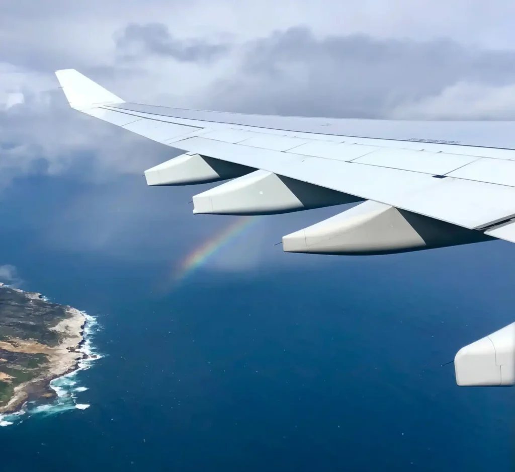 Luftaufnahme aus einem Flugzeugfenster, die einen Flügel über einer Küstenlandschaft mit einem sichtbaren Regenbogen zeigt, der sich über dem Ozean wölbt, und einer Segelyacht, die darunter unter einem bewölkten Himmel segelt.