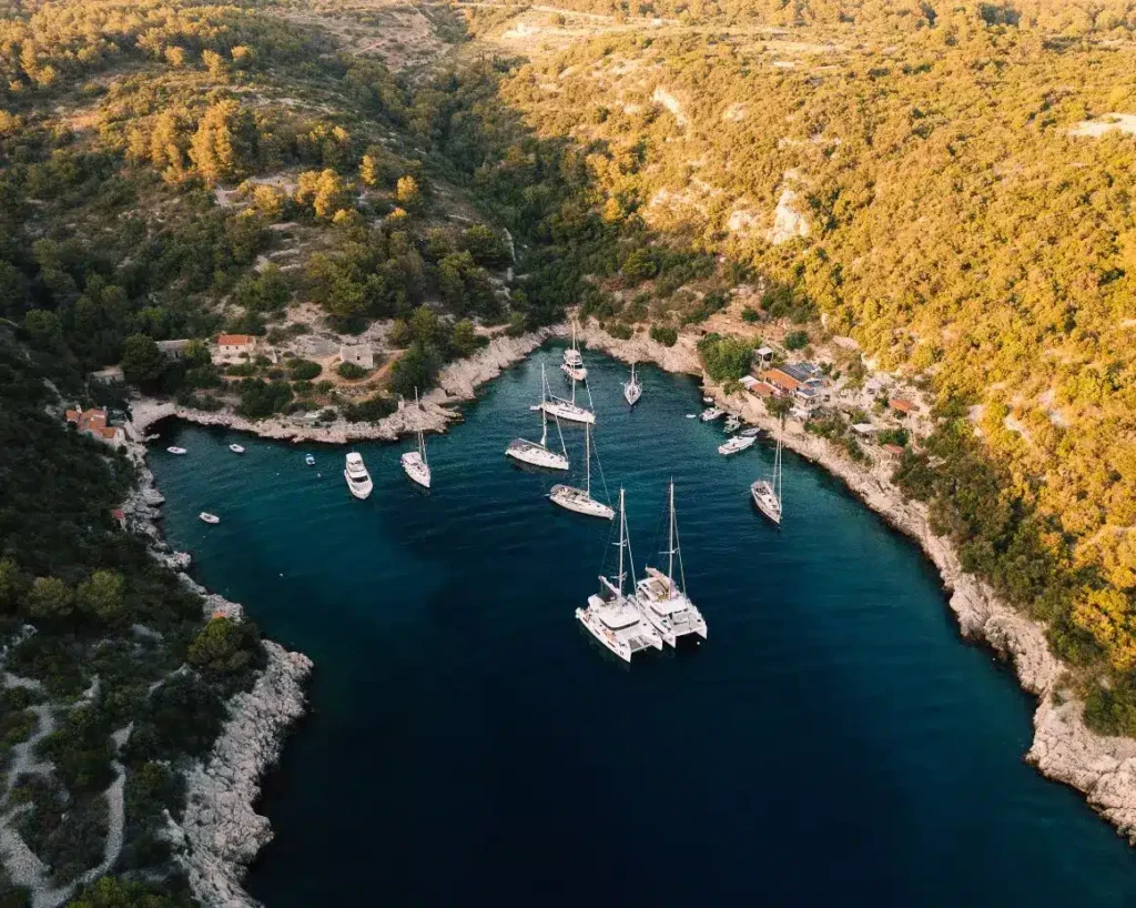 Luftaufnahme einer ruhigen Bucht mit Segelyachten, die im klaren blauen Wasser vertäut sind, umgeben von einer zerklüfteten Küste und dichten grünen Wäldern.