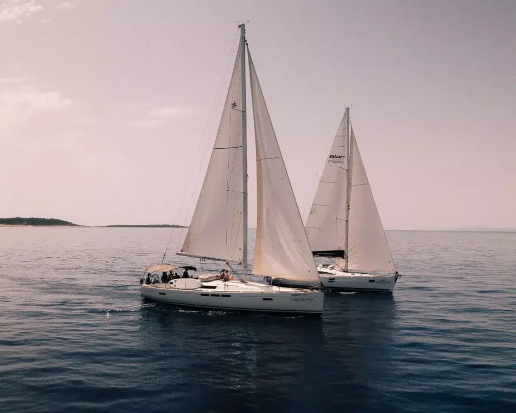 Zwei Segelyachten segeln Seite an Seite auf ruhigem, blauem Wasser mit klarem Himmel darüber. Am Heck des nächsten Segelboots ist sein Name 
