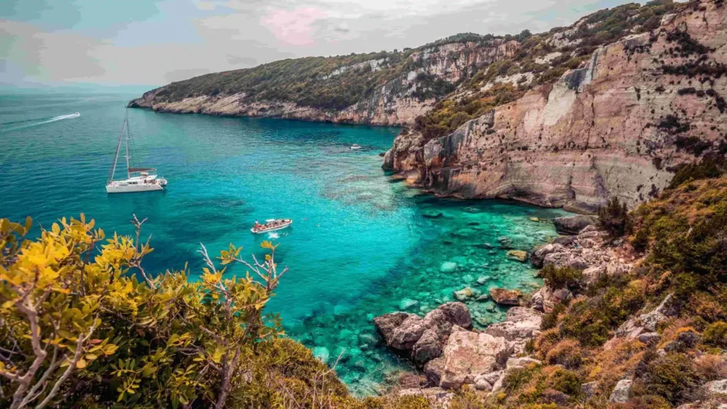 Ein malerischer Blick auf eine türkisfarbene Bucht, umgeben von üppig grünen Klippen, mit einem Segelboot und einem kleineren Boot, die während eines Segelurlaubs auf dem klaren Wasser treiben.