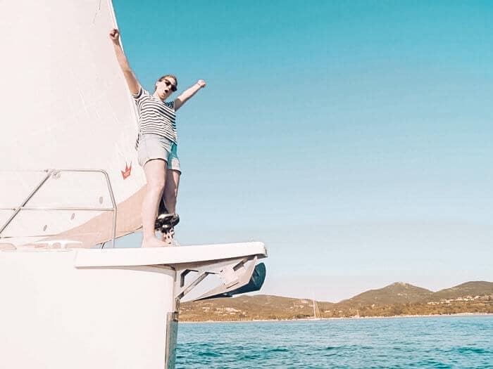 Eine Person in einem gestreiften Tanktop und Shorts steht triumphierend mit erhobenem Arm auf dem Bug einer Segelyacht, im Hintergrund das blaue Meer und Hügel.