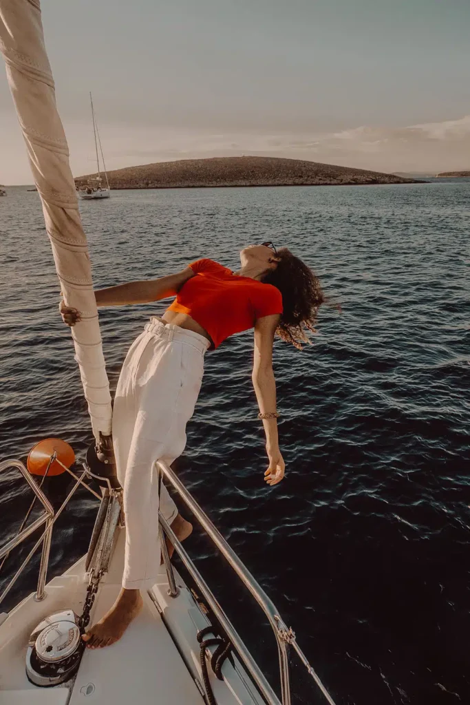 Eine Frau in einem roten Oberteil und weißen Hosen lehnt sich anmutig auf dem Bug eines Segelboots zurück und hält sich an einer senkrechten Stange fest. Umgeben von einem ruhigen blauen Meer, im Hintergrund sind Hügel unter einem teilweise bewölkten Himmel zu sehen. Die Szene auf dieser eleganten Segelyacht wirkt sowohl entspannt als auch heiter.