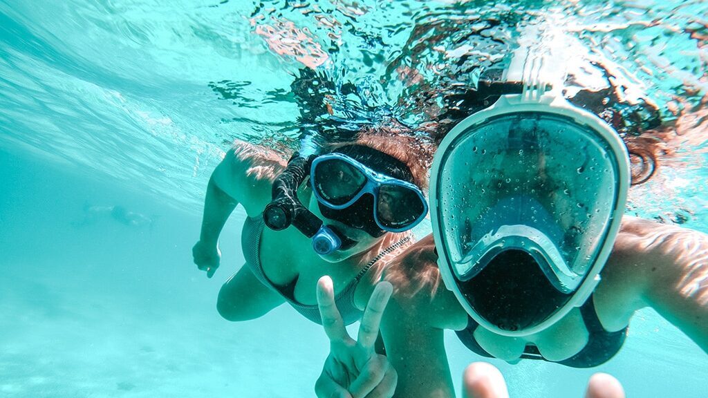Zwei Personen unter Wasser mit Schnorchelmasken machen während ihres Segelurlaubs ein Selfie. Eine Person hält mit den Fingern ein Peace-Zeichen hoch. Sie sind umgeben von klarem blauem Wasser in der Nähe ihres Katamarans.