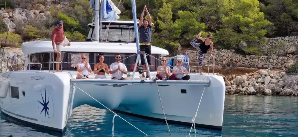 Eine Gruppe von Menschen vergnügt sich auf einer Segelyacht, die in der Nähe einer felsigen Küste vor Anker liegt. Einige sitzen, andere stehen oder posieren, alle wirken fröhlich unter dem sonnigen Himmel.