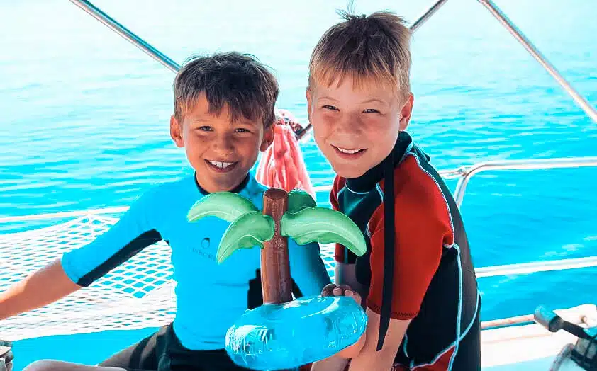 Zwei junge Jungen lächeln auf einer Segelyacht, halten ein Spielzeug mit Delfinfiguren in den Händen und stehen in Badebekleidung im hellen Sonnenlicht.