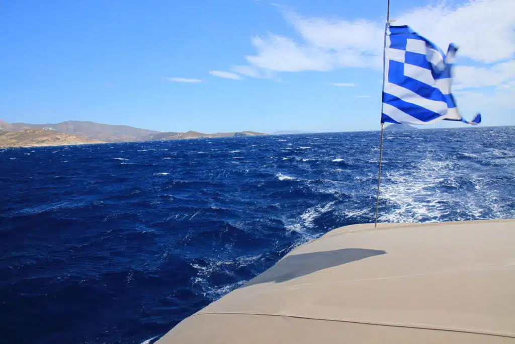Blick von einem Boot während eines Segeltörns. Zeigt eine wehende griechische Flagge am Heck mit einem leuchtend blauen Meer und Hügeln im Hintergrund unter einem klaren Himmel.