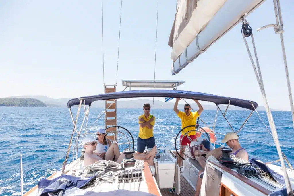 Eine Gruppe von Freunden genießt einen sonnigen Tag auf einer Segelyacht, wobei zwei Männer am Steuer stehen und die anderen sitzen, umgeben von blauem Meer und klarem Himmel.