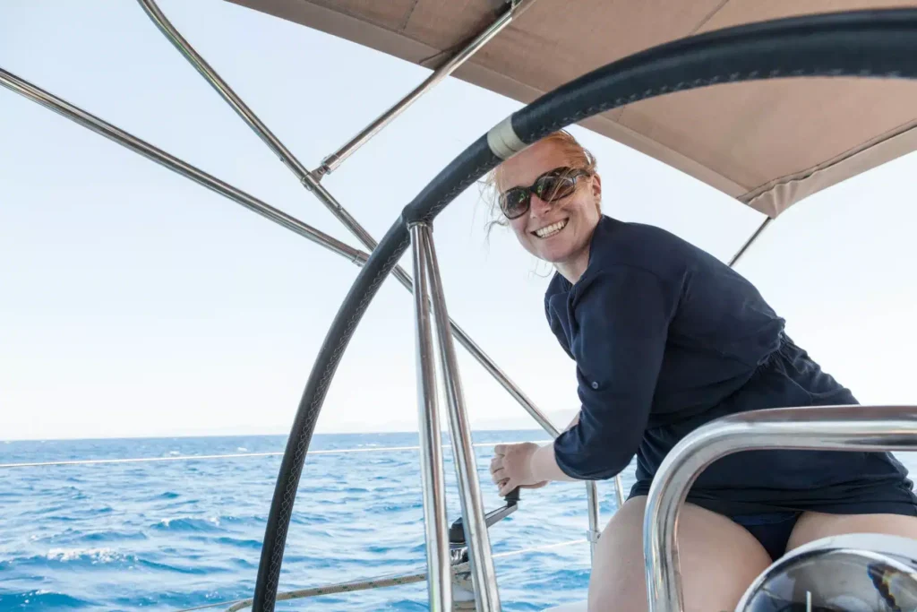 Eine Frau mit Sonnenbrille und blauem Oberteil lächelt fröhlich, während sie an einem hellen, sonnigen Tag mit blauem Himmel und ruhiger See eine Segelyacht steuert.