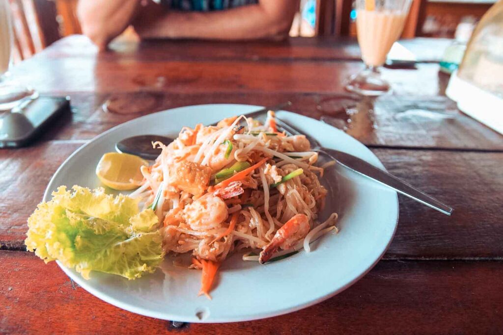 Ein Teller Pad Thai mit Garnelen, Sojasprossen und einer Limettenscheibe, serviert auf einem Holztisch mit einer Person und einem Milchshake im Hintergrund während einer Segelreise.