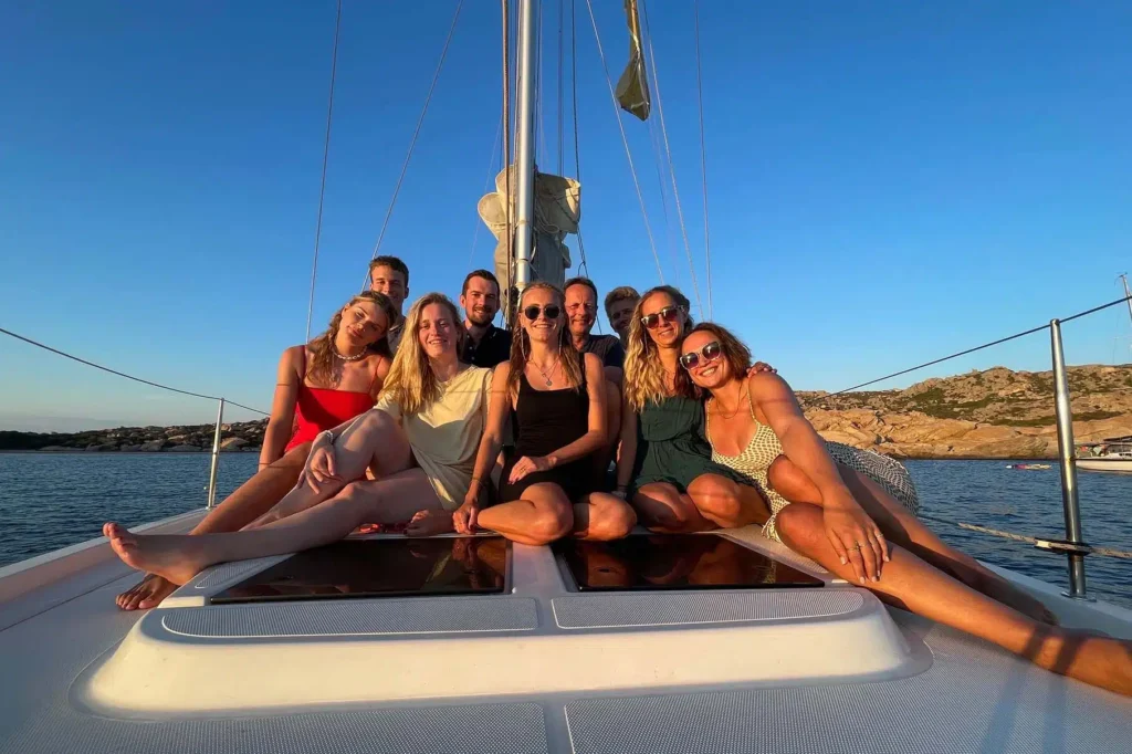 Eine Gruppe von acht Freunden posiert während ihrer Segelreise bei Sonnenuntergang auf dem Vorderdeck eines Segelboots und lächelt freudig vor der malerischen Kulisse des ruhigen Meeres und der felsigen Küste.