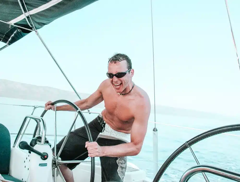 Ein fröhlicher Mann steuert eine Segelyacht, ohne Hemd, mit Sonnenbrille und einem breiten Lächeln; im Hintergrund klarer Himmel und ruhiges Meer.