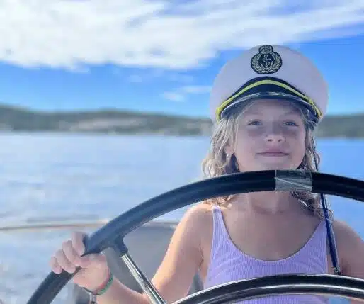 Ein junges Mädchen mit welligem blondem Haar und weißer Kapitänsmütze steht lächelnd am Steuer einer Segelyacht. In ein hellviolettes Tanktop gekleidet genießt sie während ihrer Familiensegelreise die ruhige Kulisse eines sonnenbeschienenen Sees unter einem blauen, teilweise bewölkten Himmel.