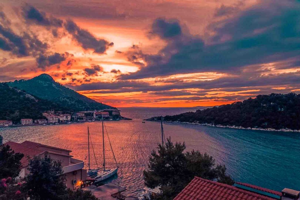 Ein malerischer Sonnenuntergang über einer Küstenstadt mit Segelbooten und einem eleganten Katamaran, der am Ufer angedockt ist. Der Himmel ist eine Mischung aus leuchtenden Orange-, Rosa- und Lilatönen, die sich im ruhigen Wasser spiegeln. Hügel und Dächer rahmen die ruhige Szene ein und schaffen eine friedliche Atmosphäre.