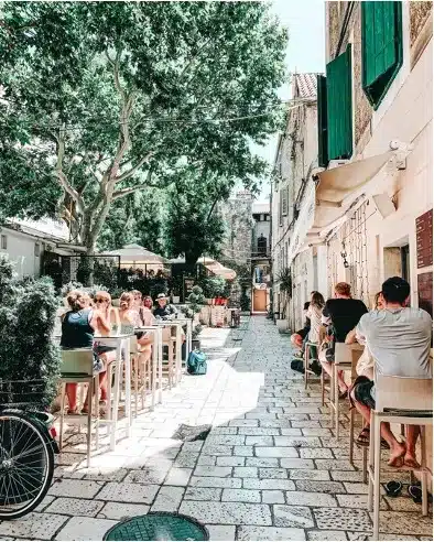 Eine belebte schmale Straße in einer europäischen Stadt mit Menschen, die in Straßencafés essen, mit Kopfsteinpflaster und üppigen grünen Bäumen, ergänzt durch Gebäude mit grünen Fensterläden. In der Nähe das Summen eines