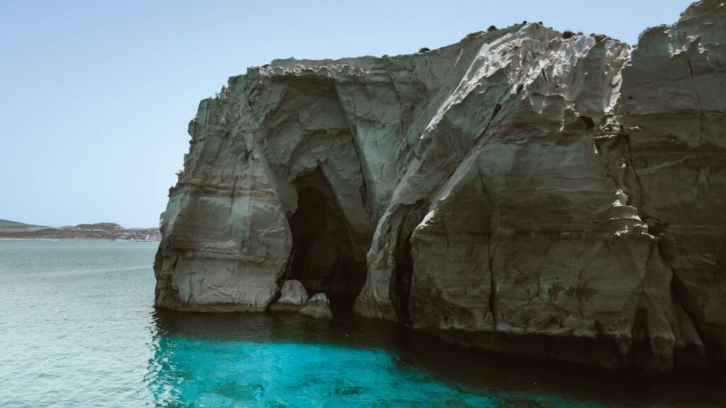Eine große Felsklippe mit einem dunklen Höhleneingang an ihrer Basis erhebt sich aus dem türkisfarbenen Wasser des Meeres. Die Felswand ist steil und zerklüftet, mit helleren und dunkleren Gesteinstönen. Der ferne Horizont zeigt einen Landstreifen unter einem klaren blauen Himmel, perfekt für Skippertraining im Mittelmeer.