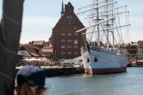 Eine Person beobachtet an einem sonnigen Tag ein großes weißes Segelschiff, das in einem Hafen mit traditionellen roten Backsteingebäuden und einem Kirchturm im Hintergrund vor Anker liegt.