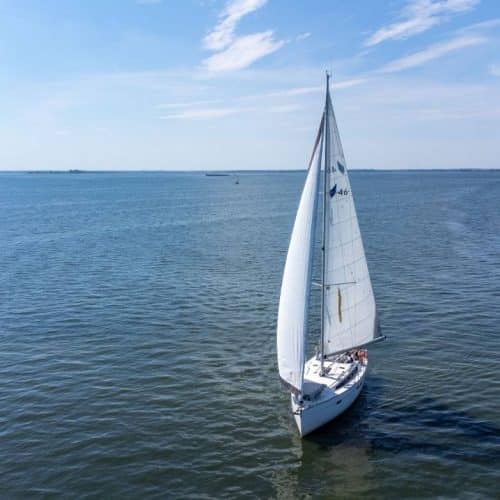 Luftbild einer weißen Segelyacht mit gesetzten Segeln, die unter einem klaren Himmel durch ruhiges blaues Wasser gleitet.