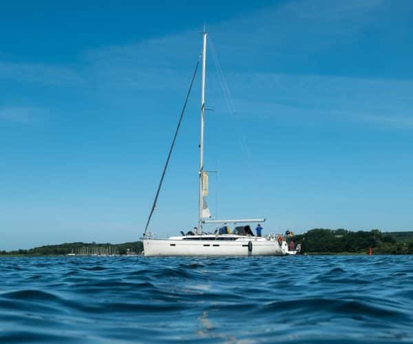 Eine Segelyacht mit weißem Rumpf und aufgerollten Segeln gleitet unter klarem Himmel auf ruhigem blauen Wasser, im Hintergrund ist eine grüne Küstenlinie sichtbar.