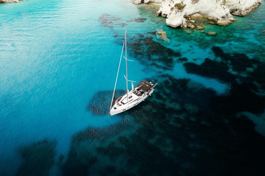 Ein Segelboot ankert in einer ruhigen, türkisfarbenen Bucht, umgeben von teilweise unter Wasser liegenden Felsen und weißen Klippen – perfekt für eine ruhige Pause während einer Atlantiküberquerung. Das Wasser variiert in Blautönen, mit Flecken von dunklerem Blau und Grün um das Boot herum.