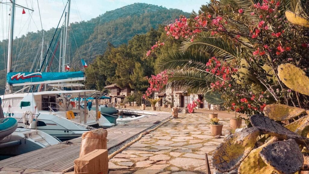 Ein malerischer Yachthafen mit Segelyachten, die entlang eines Steinwegs vertäut sind, umgeben von üppigem Grün und leuchtend rosa Blumen, mit Gebäuden am Hang im Hintergrund.