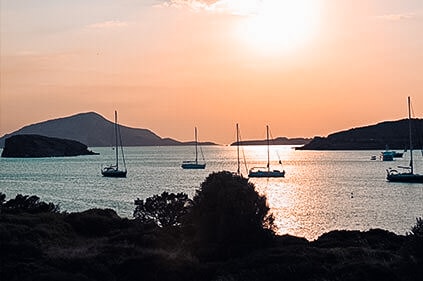 Ein heiterer Sonnenuntergang über einem ruhigen Meer mit mehreren Segelbooten, die in Küstennähe vor Anker liegen und sich vor einem pastellfarbenen Himmel mit Inseln im Hintergrund abzeichnen.