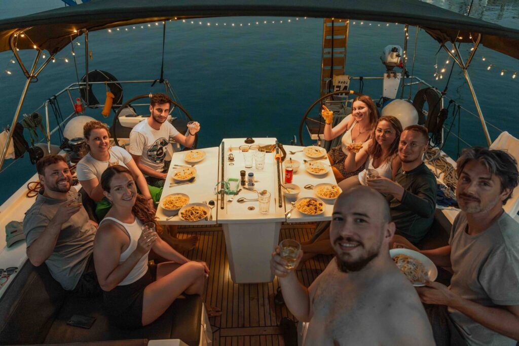 Eine Gruppe von zehn Personen sitzt auf einer Segeltour um einen Tisch und genießt gemeinsam eine Mahlzeit. Auf dem Tisch stehen Teller mit Pasta und Getränken. Die Gruppe scheint sich zu unterhalten und zu lächeln, während festliche Lichterketten eine warme Atmosphäre schaffen, während die Sonne über dem Wasser untergeht.