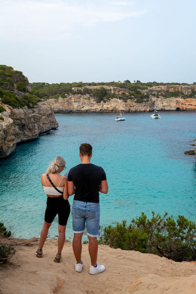 Ein Mann und eine Frau stehen auf einer Sandklippe und blicken auf eine ruhige Bucht mit klarem, türkisfarbenem Wasser. Zwei Segelboote und eine schnittige Segelyacht liegen in der Bucht vor Anker. Die Klippe ist von üppigem Grün und Felsformationen umgeben. Der Himmel ist klar mit einem Hauch von Wolken.