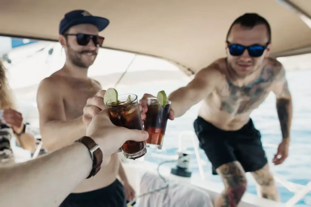 Drei Freunde stoßen mit Getränken auf einer Segelyacht an. Zu sehen sind zwei Männer, einer davon tätowiert, die einen sonnigen Tag auf dem Wasser genießen.