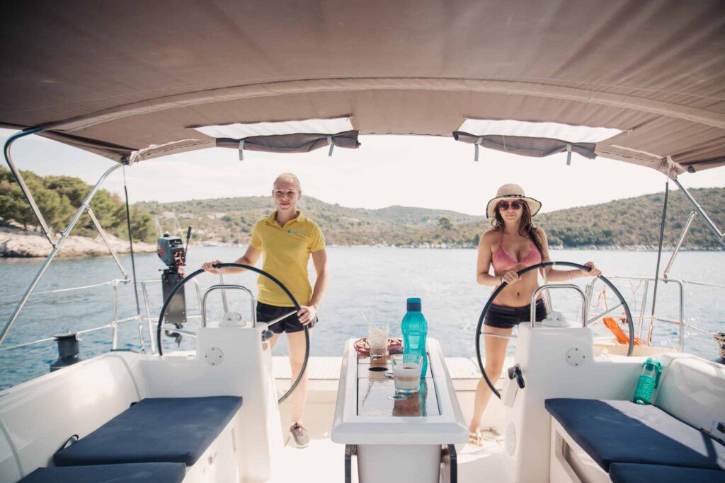 Zwei Personen, ein Mann in einem gelben Hemd und eine Frau in einem rosa Bikinioberteil und Hut, steuern einen Katamaran im hellen Sonnenlicht. Sie stehen an Doppelsteuerrädern auf beiden Seiten des Cockpits des Bootes, zwischen ihnen ein Tisch und nautische Ausrüstung. Im Hintergrund ist eine malerische Aussicht auf Wasser und Hügel zu sehen, die ihren perfekten Segeltörn verkörpert.