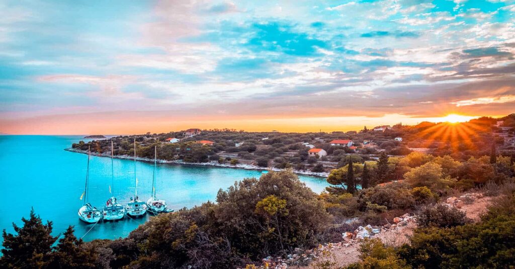 Ein Panoramablick auf eine ruhige Küstenlandschaft bei Sonnenuntergang, mit einer kleinen Bucht mit Booten, ideal für einen Segelurlaub, umgeben von üppigem Grün und Häusern unter einem strahlenden Himmel.