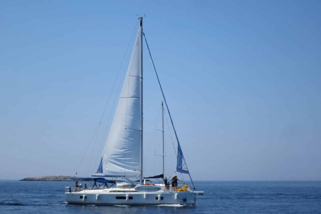 Ein Katamaran mit gesetzten Segeln segelt auf ruhiger See unter einem klaren blauen Himmel, perfekt für einen Segelurlaub, mit ein paar Leuten auf dem Deck.