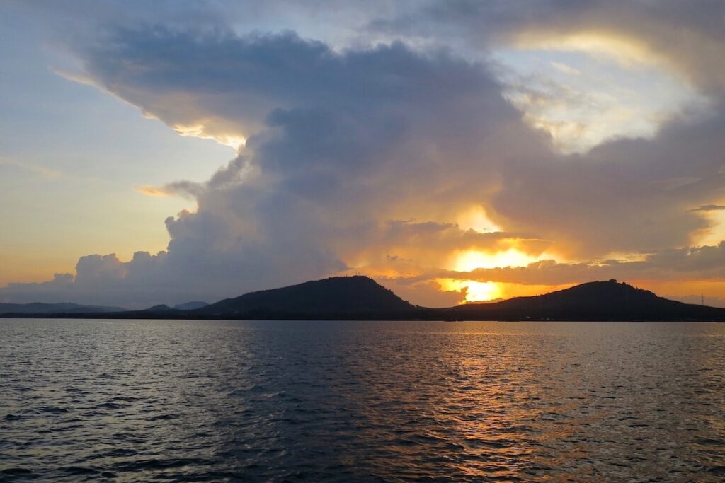Ein leuchtender Sonnenuntergang über einem ruhigen Meer mit einer großen Wolkenformation darüber und Hügelsilhouetten am Horizont. Der Himmel ist eine Mischung aus Orange- und Blautönen, vom Deck einer Segelyacht aus betrachtet.