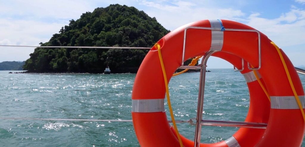 Ein orangefarbener Rettungsring, der an der Reling einer Segelyacht befestigt ist, mit einer üppig grünen Insel und dem blauen Meer im Hintergrund unter einem teilweise bewölkten Himmel.