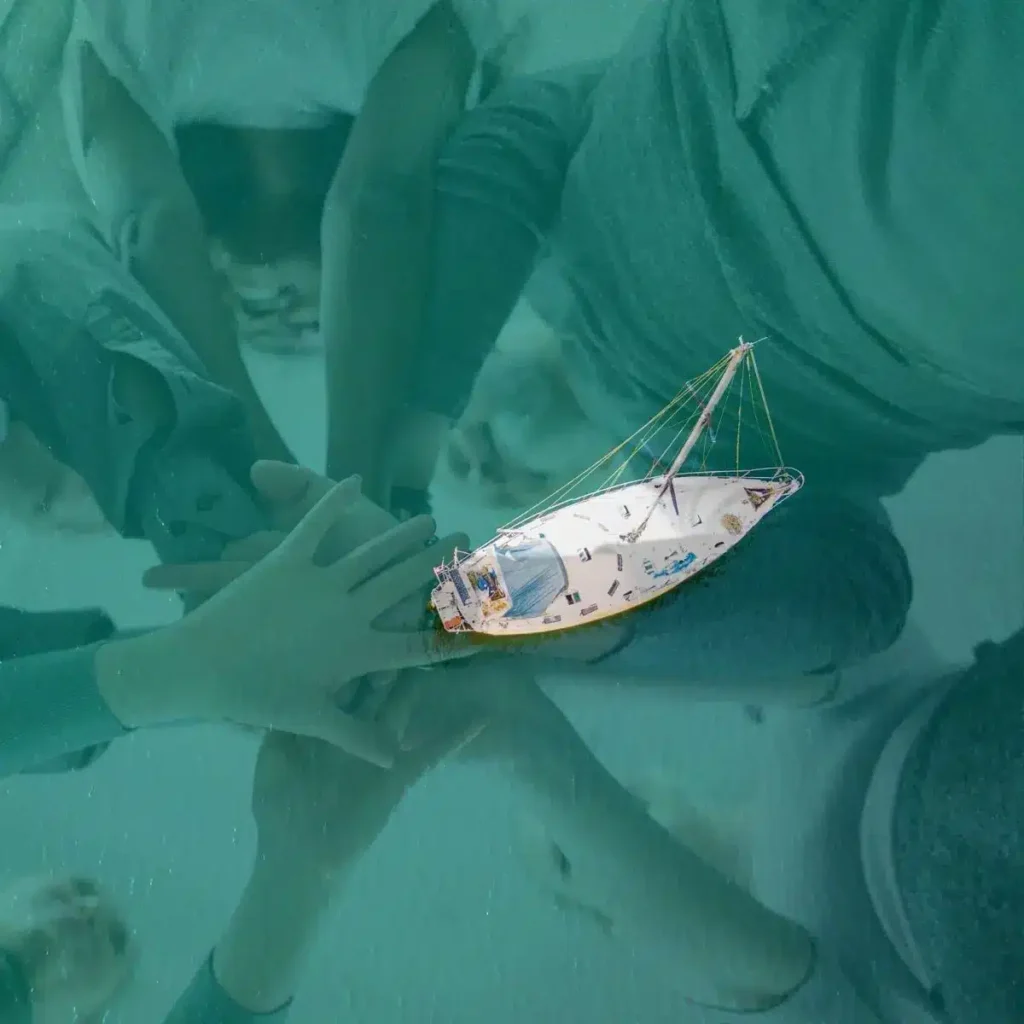 Ein Miniatursegelboot schwimmt auf klarem, türkisfarbenem Wasser über einer Unterwasserszene mit sandigem Meeresboden und Schatten von Menschen, die während eines Segeltörns vom Sonnenlicht geworfen werden.