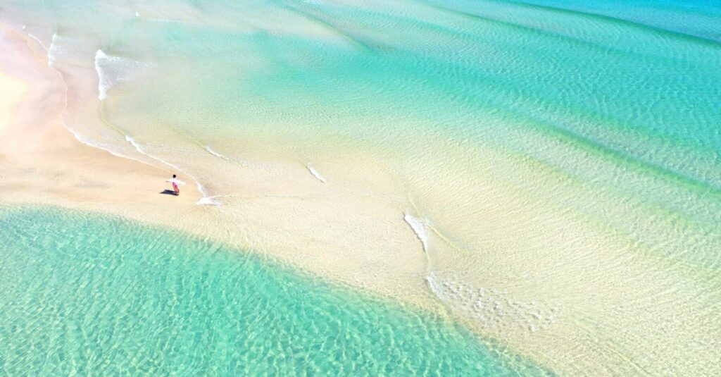 Luftaufnahme einer Person, die an einem unberührten Sandstrand entlangspaziert, an dessen Ufer kristallklares, türkisfarbenes Wasser sanft plätschert. Die Szene fängt die ruhige und weite Schönheit des Strandes ein und weckt Gedanken an eine Segelreise.