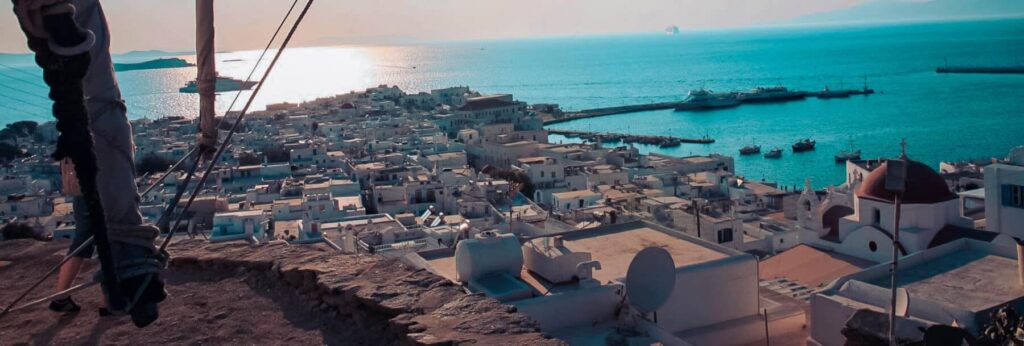 Ein Panoramablick auf eine griechische Inselstadt in der Abenddämmerung mit weißen Gebäuden und Kirchen mit blauen Kuppeln, mit dem Meer und Katamaranen im Hintergrund.