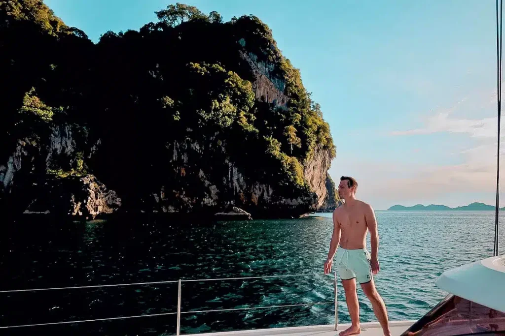 Eine Person in Shorts steht auf einer Jacht und segelt unter klarem Himmel entlang der tropischen Küste mit üppigen Klippen und ruhigem blauen Wasser.