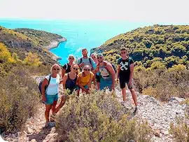 Eine Gruppe von sieben Personen posiert während ihres Segelurlaubs auf einem Wanderweg mit üppigem Grün und einem malerischen Flussblick im Hintergrund unter einem strahlend blauen Himmel.