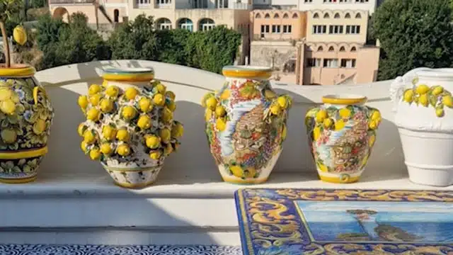 Bunte, aufwendig verzierte Keramikvasen mit gelben Zitronen, die auf einem Sims angeordnet sind. Im Hintergrund sind Gebäude und Grünflächen zu sehen. Eine Vase ist reich mit dreidimensionalen Zitronenverzierungen verziert, während die anderen mit malerischen Motiven bemalt sind, die an einen ruhigen Tag beim Mitsegeln auf einer Segelyacht erinnern.